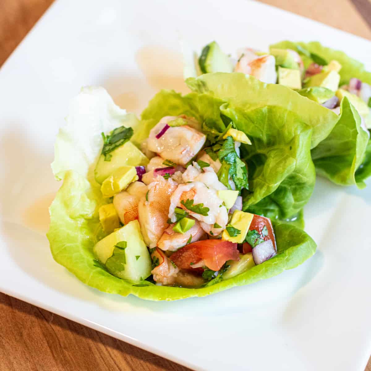 Lettuce wraps with shrimp ceviche, diced red onion, cucumber, avocado, tomato, and cilantro on a white square plate.