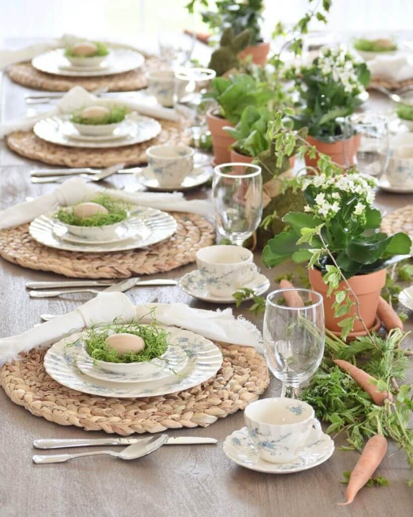 Natural woven chargers with blue and white dishes. Small bowl at each place setting is filled with Easter grass and an egg. Down the center of the table are white flowers and greenery in clay pots and carrots with green stems.