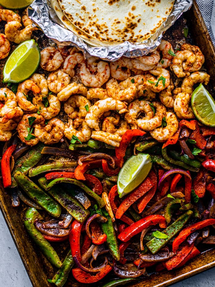 Sheet pan with cooked peppers, shrimp, tortillas and lemon wedges.